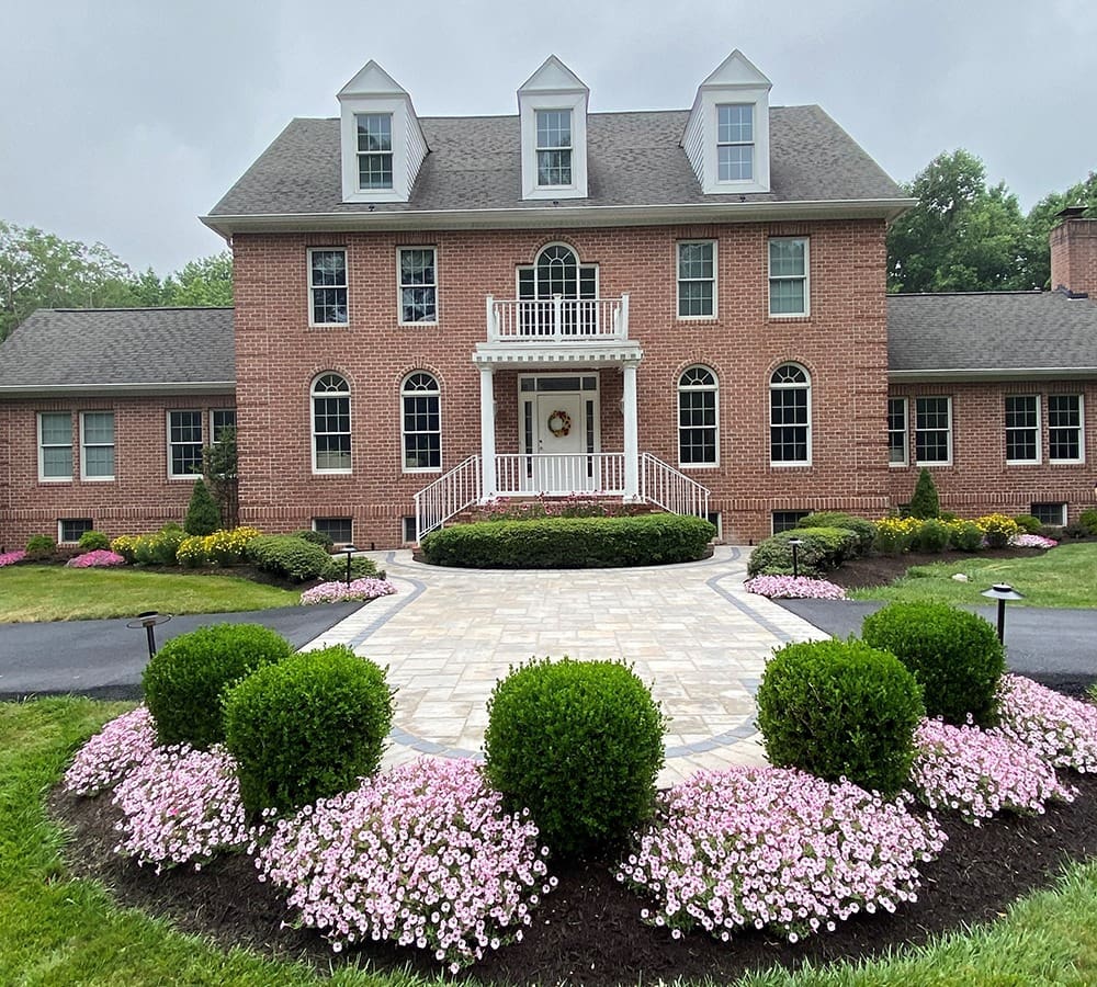 A large red-brick house with a symmetrical design and three dormer windows on the roof. The front features a white-pillared entrance and a balcony above. A paved walkway, surrounded by neatly trimmed hedges and pink flowers, leads to the front door.